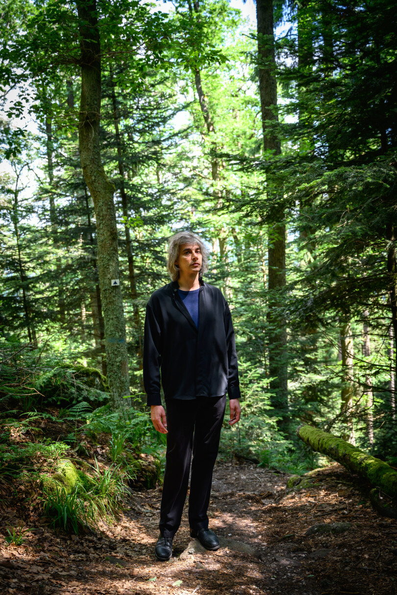 Le comédien Micha Lescot, en noir et bleu, se détache sur le vert des arbres et le brun des feuilles mortes.